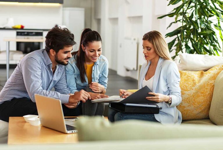 Remote worker at a home desk with a laptop, monitor, office files, and household items nearby.